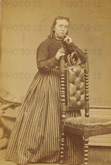 Portrait of an unknown woman from the Marmelstein family, photograph, carte-de-visite, photographer: Caspel & Stapert, S. van, Amsterdam, 1850 - 1900, paper, pasteboard, albumen print, albumendruk, height 111 mm x width 65 mm, historical persons, adult woman, A sepia-toned portrait of a person standing beside an ornate wooden chair with a tufted leather back and seat, the person wearing a dark long-sleeved bodice and a long skirt with vertical stripes; one hand rests on the turned finial of the chair while the other hand is raised near the face, and the person looks slightly forward with a neutral expression, hair pulled back and styled; the background is plain and textured.