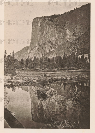 El Capitan, Yosemite Valley. 3, 300 feet high, View of El Capitan in the Yosemite Valley, page, photomechanical print, Yosemite Valley, c. 1866 - in or before 1876, North America, California, paper, photoaquatint, height 157 mm x width 108 mm, Print opposite title page., rock-formations, A vertical monochrome photograph showing a steep rocky cliff rising from a forested foothill, with rugged rock faces and textured slopes; a line of pine trees stands at the base of the cliff, and a grassy meadow lies between the trees and a calm reflective body of water in the foreground that mirrors the cliff and some trees; the sky above is clear and pale, and the image has a grainy, aged paper appearance with a narrow light border around it.