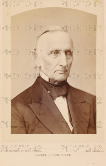 Samuel S. Schmucker, photograph, page, photographer: William H. Tipton, Gettysburg, c. 1877 - in or before 1882, North America, Pennsylvania, paper, albumen print, height 148 mm x width 103 mm, Person, Samuel Simon Schmucker, adult man, A sepia portrait of an older man shown from the chest up wearing a dark suit jacket, white shirt, and a dark bow tied at the collar, with short combed hair, prominent sideburns, and a lined face with a neutral expression, set against a plain light background and framed by a light border.
