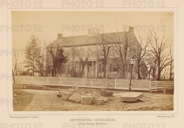 Gettysburg grammar school, photograph, page, photographer: William H. Tipton, Gettysburg, c. 1877 - in or before 1882, North America, Pennsylvania, paper, albumen print, height 102 mm x width 144 mm, facade (of house or building), university building, college, A sepia-toned photograph of a two-story brick building behind a picket fence with bare trees in front of it, a pitched roof with chimneys and a small railed structure on top, shuttered windows and a central doorway with a small porch, a dirt road and scattered large stones in the foreground, a slightly leaning street lamp near the fence, and additional trees and fencing visible along the sides.