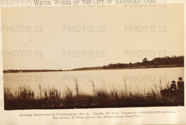 Water works of the city of Hartford, Conn, Storage reservoir at West Hartford (no. 4). Depth, 20 feet. Capacity 600, 000, 000 gallons. Elevation, 27 feet above Connecticut river, photograph, page, Connecticut, c. 1883 - in or before 1884, photographer, North America, paper, albumen print, height 108 mm x width 180 mm, water course, A wide sepia-toned photograph showing a calm body of water extending toward a distant tree-lined shoreline, with tall grasses and reeds in the foreground along the near bank; on the right edge two seated figures wearing dark clothing and hats are partially visible among the grasses; the sky above the water is mostly clear and light in tone.
