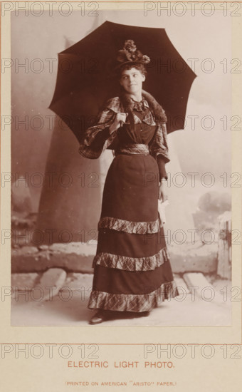 Electric Light Photo, Portrait of an unknown woman with an umbrella, photograph, page, inscription: 'Pinted on American 'Aristo' Paper', c. 1888 - in or before 1893, photographer, paper, daylight collodion silver print, height 140 mm x width 98 mm, Photo two pages before page 178., adult woman, A sepia-toned portrait of a woman standing and holding a large dark umbrella over her shoulder, wearing a long dark dress with patterned trim bands on the skirt and matching patterned sleeves, a fur or furlike collar, and a decorative hat with flowers, her hair styled up and her face turned slightly to one side with a faint smile, one hand grasping the umbrella handle and the other holding a light-colored cloth, with a studio backdrop and a visible floor area beneath her feet.