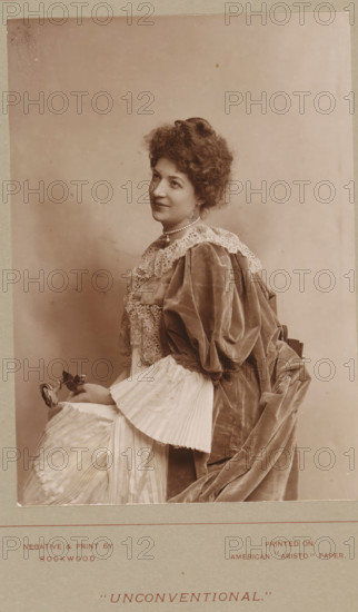 Unconventional, Portrait of an unknown woman with a rose in her hand, photograph, page, inscription: 'Printed on American 'Aristo' paper', photographer: Rockwood, c. 1888 - in or before 1893, paper, daylight collodion silver print, height 140 mm x width 100 mm, Photo two pages before page 382., historical persons, flowers, adult woman, A sepia-toned portrait of a seated woman shown in profile from the waist up, wearing a voluminous dress with a lace collar and puffed velvet sleeves, a pleated fabric cuff visible at her wrist, and a layered skirt; she has curly hair swept up and wears a short strand of pearls at her neck, her head slightly tilted and her gaze directed upward; one hand rests on her lap holding a small flower, and the draped fabric of her dress falls in soft folds behind her against a plain background.