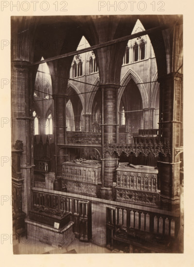 Chapel of St. Edmund at Westminster Abbey, photograph, page, photographer: John Harrington, c. 1864 - in or before 1869, paper, albumen print, height 194 mm x width 139 mm, interior of church, abbey, monastery, convent, Roman Catholic church, parts of church interior: chapel, London, Westminster Abbey, The image shows the interior of a large stone building with tall, ribbed pointed arches and clustered columns supporting vaulted ceilings; ornate carved stone screens and tombs with recumbent effigies sit within the space, and a decorative railing and intricate sculpted panels are visible in the foreground; light streams through tall arched windows in the background, casting soft highlights on the stone surfaces and emphasizing the vertical architectural elements.