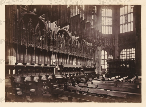 Chapel of Henry VII at Westminster Abbey, photograph, page, photographer: John Harrington, c. 1864 - in or before 1869, paper, albumen print, height 139 mm x width 195 mm, interior of church, abbey, monastery, convent, Roman Catholic church, parts of church interior: chapel, London, Westminster Abbey, A sepia-toned interior photograph showing rows of wooden pews in the foreground leading toward ornately carved choir stalls along the left side, the stalls featuring tall, decorative wooden panels with intricate tracery and sculpted figures. Fabric banners hang from above, draping down into the middle of the image. Large, tall windows with mullioned panes occupy the background on the right, letting bright light stream into the space and casting patterned highlights. Stone arches and detailed woodwork frame openings and an elevated wooden structure with additional carvings appears toward the right rear. The overall scene emphasizes the interplay of dark wood, carved ornamentation, hanging ...