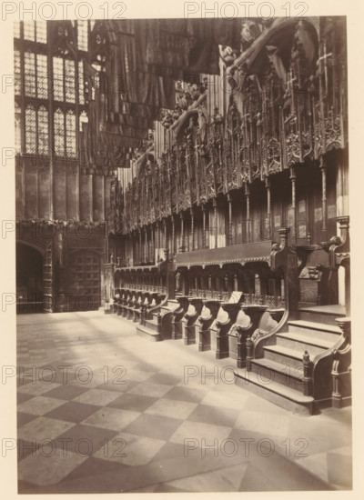 Chapel of Henry VII at Westminster Abbey, photograph, page, photographer: John Harrington, c. 1864 - in or before 1869, paper, albumen print, height 177 mm x width 125 mm, interior of church, abbey, monastery, convent, Roman Catholic church, parts of church interior: chapel, London, Westminster Abbey, A sepia-toned interior photograph showing a long row of ornately carved wooden choir stalls with high backs and armrests, a stepped platform at one end, and decorative panels and vertical columns above them; a patterned stone floor of alternating light and dark squares stretches toward the foreground; tall leaded glass windows with geometric panes occupy the left wall, and heavy hanging textiles or banners are visible near the ceiling; stone walls and an archway with a large wooden door appear at the far left, and detailed woodwork and tracery fill the upper right area of the image.