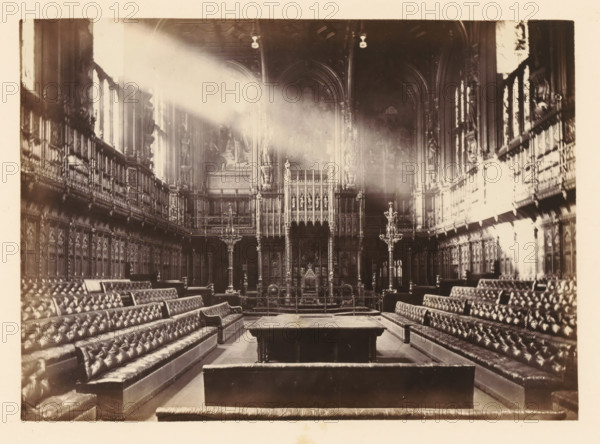 View of the interior of the Commons Chamber, photograph, page, photographer: John Harrington, London, c. 1864 - in or before 1869, paper, albumen print, height 139 mm x width 195 mm, palace, interior, representation of a building, Houses of Parliament, A wide interior view of a grand hall with high vaulted ceilings and tall windows along both side walls, sunlight streaming in as a bright diagonal beam. The room contains long, upholstered benches arranged in parallel rows facing a raised ornate screen at the far end, which features intricate carved panels, statues, and decorative columns. Two tall, ornate candle stands flank the central aisle near the screen. The side walls are lined with elaborate wood paneling and carved figures set into niches above the panels. The ceiling shows decorative moulding and hanging light fixtures. The overall tone of the image is sepia, and the floor appears smooth and reflective in places.