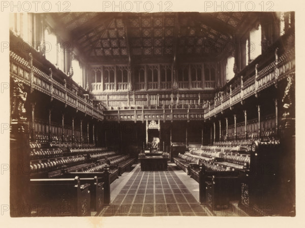 Interior of House of Commons, photograph, page, photographer: John Harrington, London, c. 1864 - in or before 1869, paper, albumen print, height 138 mm x width 194 mm, palace - AA - civic architecture: inside, Houses of Parliament, A large ornate hall with high, patterned ceiling and tall windows along both sides, flanked by two long rows of carved wooden benches with upholstered seats and decorative armrests, a central aisle with a tiled floor leading to a raised platform area at the far end where a large, elaborately carved chair or throne sits beneath an ornate canopy, intricate wood paneling and balustrades lining the upper galleries on both sides, sculpted figures and decorative columns integrated into the woodwork, and the scene rendered in a sepia-toned photograph.