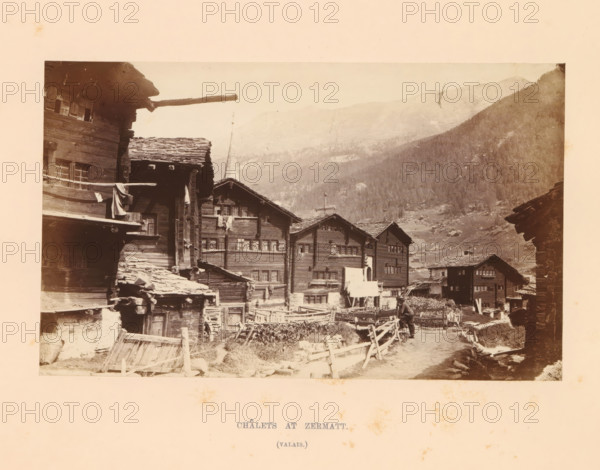 View of chalets in Zermatt, Châlets at Zermatt, photograph, page, inscription: '(valais)', photographer: Stephen Thompson, Zermatt, c. 1863 - in or before 1868, paper, albumen print, height 107 mm x width 176 mm, street (city(-scape) with figures, staffage), hut, cabin, lodge, A sepia-toned photograph showing a row of wooden chalets with steep shingled roofs and multiple small windows, built close together along a dirt path lined with simple wooden fences; a person is standing on the path near the foreground beside the fences; some cloth is hanging from a balcony or railing on one of the buildings; a tall spire rises behind the chalets and tree-covered mountains form the background under a pale sky.