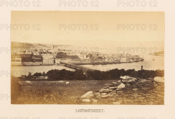 View of Londonderry, Londonderry, photograph, page, photographer: John Hudson, c. 1862 - in or before 1867, paper, albumen print, height 108 mm x width 177 mm, city-view in general, 'veduta', landscapes with waters, waterscapes, seascapes (in the temperate zone), bridge, A sepia-toned panoramic view showing a riverside town with a long bridge crossing the water, buildings and warehouses lining the near shore, a tall church spire rising among the rooftops, several ships and boats moored along the waterfront, tree-covered areas and open grassy land in the foreground, a rocky stone wall and scattered boulders at the bottom edge, distant rolling hills across the river, and a tall cylindrical chimney standing near the water.