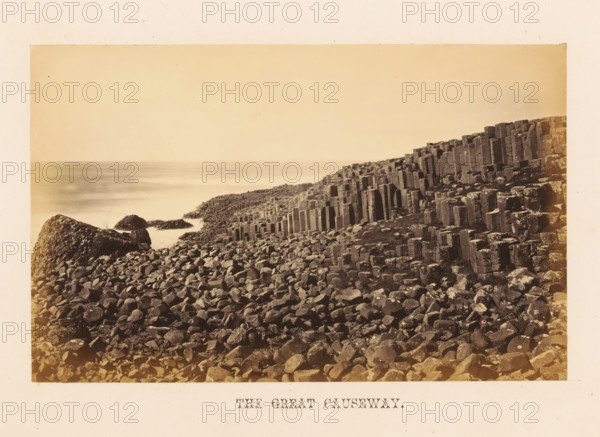 The Great Causeway, View of Causeway, photograph, page, photographer: John Hudson, Giant's Causeway, c. 1862 - in or before 1867, John Hudson, photographer, Causeway Coast and Glens, paper, albumen print, height 110 mm x width 176 mm, landscapes with waters, waterscapes, seascapes (in the temperate zone), rock-formations, sea, A coastal scene showing a rocky shoreline with numerous dark, irregular stones in the foreground and clustered, column-like basalt formations rising toward the right, a few larger rounded boulders near the water, a calm sea stretching into the distance under a pale sky, and a narrow light border framing the image.