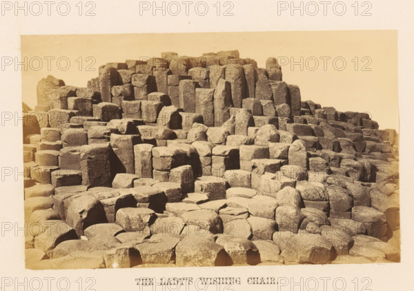 View of rock formation the Lady's Wishing Chair, The Lady's Wishing Chair, photograph, page, photographer: John Hudson, Giant's Causeway, c. 1862 - in or before 1867, John Hudson, photographer, Causeway Coast and Glens, paper, albumen print, height 110 mm x width 178 mm, landscapes, rock-formations, A sepia-toned photograph showing a large area of interlocking, columnar rock formations with flat tops and vertical sides, arranged in terraces and clusters that rise toward the back of the image; the rocks display rough, weathered surfaces and edges with visible cracks and joints, and varying heights and shapes creating a stepped, geometric pattern across the scene, with a pale, empty sky above and a narrow pale border around the photograph.