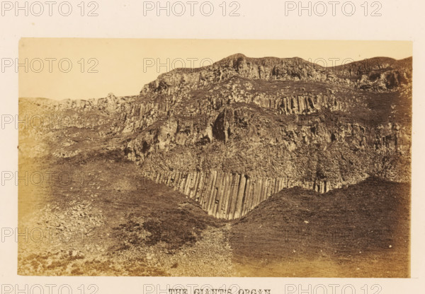 The Giant's Organ, View of rock formation The Giant's Organ, photograph, page, photographer: John Hudson, Giant's Causeway, c. 1862 - in or before 1867, John Hudson, photographer, Causeway Coast and Glens, paper, albumen print, height 109 mm x width 180 mm, landscapes, rock-formations, A sepia-toned photograph of a rocky hillside with layered, column-like rock formations and rough textured slopes, showing vertical striations and rugged cliff faces, with patches of sparse vegetation and scattered stones across the uneven ground, framed by a pale border.
