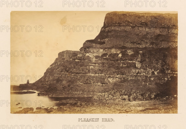 Pleskin Head, View of Pleskin Head rock formation, photograph, page, photographer: John Hudson, Giant's Causeway, c. 1862 - in or before 1867, John Hudson, photographer, Causeway Coast and Glens, paper, albumen print, height 109 mm x width 177 mm, landscapes with waters, waterscapes, seascapes (in the temperate zone), rock-formations, A sepia-toned photograph showing a rugged coastal cliff rising steeply from a rocky shoreline, with layered rock formations and vertical columnar textures on the upper face; the foreground contains a shoreline of scattered rocks and pebbles leading to calm water that laps against the base of the cliff, and a distant rock outcrop projecting into the water at the left edge; the sky above is pale and mostly empty.