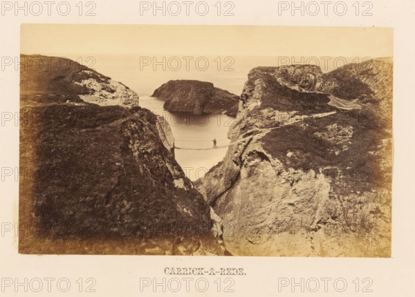 View of an unknown man on the Carrick-a-Rede rope bridge, Carrick-a-Rede, photograph, page, photographer: John Hudson, Ballintoy, c. 1862 - in or before 1867, Causeway Coast and Glens, paper, albumen print, height 110 mm x width 179 mm, landscapes with waters, waterscapes, seascapes (in the temperate zone), suspension bridge, A sepia-toned photograph showing a narrow rope bridge suspended between two rocky cliffs over a calm body of water, with a solitary figure standing on the bridge; steep rocky shores and grassy ledges flank the inlet, and rocky islands or outcrops rise in the distance under a pale sky.