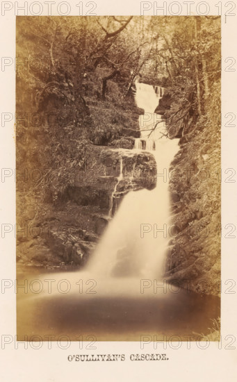 O'Sullivan's Cascade, View of O'Sullivan's Waterfall, photograph, page, photographer: John Hudson, Killarney, c. 1862 - in or before 1867, paper, albumen print, height 174 mm x width 108 mm, waterfall, Killarney National Park, A vertically oriented sepia photograph showing a multi-tiered waterfall cascading down rocky ledges surrounded by dense foliage and trees, with water flowing from an upper narrow drop into wider falls and continuing into a smooth misty pool at the base; rock faces and grassy vegetation frame both sides of the falls, and branches and tree trunks are visible among the foliage.