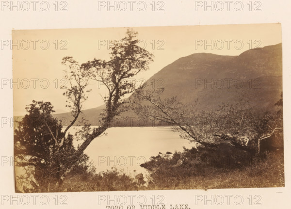 Torc or Middel Lake, View of Middel Lake, photograph, page, photographer: John Hudson, Killarney, c. 1862 - in or before 1867, paper, albumen print, height 108 mm x width 178 mm, lake, landscapes with waters, waterscapes, seascapes (in the temperate zone), mountains, Killarney National Park, A sepia-toned landscape photograph showing a body of water bordered by rugged shoreline and low vegetation, with tree branches arching over the water from both sides; a distant mountain rises behind the water, its slopes textured with rock and vegetation, and the sky above is pale and empty.