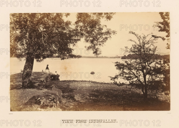 View from Innisfallen, View from Innisfallen with an unknown woman by the water, photograph, page, photographer: John Hudson, Killarney, c. 1862 - in or before 1867, paper, albumen print, height 108 mm x width 176 mm, adult man, landscapes with waters, waterscapes, seascapes (in the temperate zone), Innisfallen, A sepia-toned lakeside scene framed by large trees with overhanging branches, showing a stony shoreline in the foreground and a calm body of water extending to a distant tree-lined horizon, with a small rowboat pulled up near the shore and a woman in a long dress standing close to it, a child seated nearby, a solitary rock protruding from the water, and scattered rocks and foliage along the shore.
