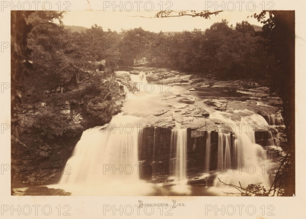 View of Bonnington Linn Waterfall, Bonnington Linn, photograph, page, South Lanarkshire, c. 1862 - in or before 1867, photographer, paper, albumen print, height 116 mm x width 178 mm, landscapes with waters, waterscapes, seascapes (in the temperate zone), waterfall, A sepia-toned photograph of a wide waterfall cascading over layered rock ledges with smooth, silky water flow; surrounding the falls are rocky banks and dense trees and foliage on both sides, with branches and leaves framing the top edge; flat rock shelves and irregular stones are visible in the river above the main drop, and mist and flowing water fill the lower foreground.