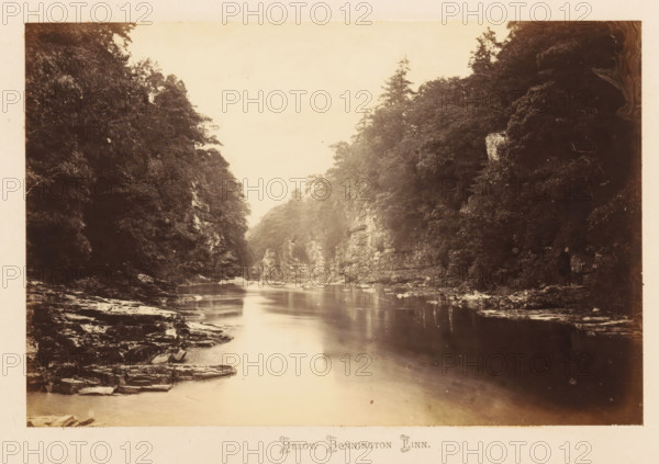 View of the Clyde below the Bonnington Linn, Below Bonnington Linn, photograph, page, South Lanarkshire, c. 1862 - in or before 1867, photographer, paper, albumen print, height 116 mm x width 179 mm, river, landscapes with waters, waterscapes, seascapes (in the temperate zone), A sepia-toned landscape photograph showing a calm river flowing between steep, rocky banks lined with dense trees and foliage; the river surface reflects light and the surrounding cliffs, with flat rock ledges and scattered boulders visible along the water’s edge, and tall trees rising above the cliffs on both sides under a pale sky.