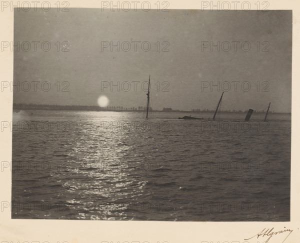 View of a sinking ship with a setting or rising sun, photograph, page, signature: 'Algrain', photographer: Alfred Algrain, c. 1885 - in or before 1890, paper, platinum process, height 158 mm x width 118 mm, landscapes with waters, waterscapes, seascapes (in the temperate zone), ships (in general), sun as celestial body, A wide expanse of water fills the foreground with rippling reflections leading toward a distant horizon, where a bright circular light sits near the waterline and casts a shimmering path across the surface; several tall, thin masts and a partially submerged structure rise from the water near the horizon, and a low, dark shoreline with a line of small vertical shapes stretches behind them under a vast, evenly toned sky.