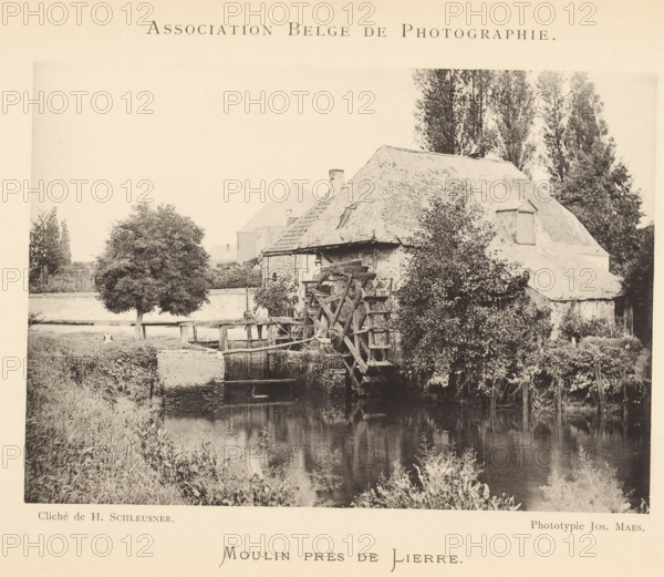 View of a watermill, Moulin près de Lierre, page, photomechanical print, inscription: 'Bulletin de l'Association belge de photographie', maker: Henri Schleusner, maker: Joseph Maes, c. 1885 - in or before 1890, Henri Schleusner, Joseph Maes, paper, collotype, photoprint, height 109 mm x width 155 mm, landscapes with waters, waterscapes, seascapes (in the temperate zone), watermill, A sepia-toned photograph shows a riverside scene with a thatched-roof building partially covered in climbing plants and foliage, a large wooden waterwheel attached to the building, a calm reflective body of water in the foreground, grassy banks and reeds along the water, a leafy tree near a simple wooden fence on the far bank, and tall trees rising behind the building.