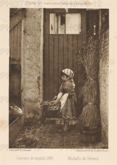 View of a girl with a basket near a goat, page, photomechanical print, inscription: 'Bulletin de l'Association belge de photographie, Concours de negatifs 1889 Medaille de Vermeil', maker: Henri Schleusner, maker: Josef Löwy, c. 1885 - in or before 1890, Henri Schleusner, Josef Löwy, paper, photoaquatint, height 152 mm x width 108 mm, girl (child between toddler and youth), goat, container made of plant material other than wood: basket, A sepia-toned photograph shows a young girl standing in front of a wooden door, wearing a striped headscarf, a shawl, a patterned skirt, and knitted socks with shoes; she holds a woven basket filled with produce and looks downward. To her right is a tall bundle of straw tied and placed in a wooden tub, and to her left a rough stone wall and a low opening where a goat's head peers out. The ground is cobbled and worn, and the scene is framed by textured walls and doorframes.