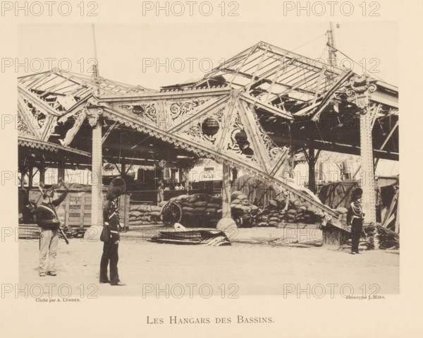 Les hangars des bassins, Sheds whose roof collapsed due to an explosion of a munitions factory in Oosterweel near Antwerp in 1889, page, photomechanical print, number: '10', photographer: Albéric Lunden, Oosterweel, cliché maker: anonymous, Belgium, printer (person): J. Maes (possibly), Antwerp, 1889, printer (person), paper, collotype, photoprint, height 165 mm x width 230 mm, Image on page 10., the soldier, the soldier's life, ruin of a building, architecture, hangar, Explosion of a munitions factory in Oosterweel in 1889, A large ornate iron structure with decorative cutouts and scalloped edges forms a partially ruined roof over a covered area filled with stacked sacks and crates, wooden beams, and a large wagon wheel; several vertical columns support the framework and ropes and rigging extend upward from the roof; three uniformed figures stand in the foreground wearing dark coats and tall hats, one holding a long object and another with a bag at his side; sections of the roof show ...