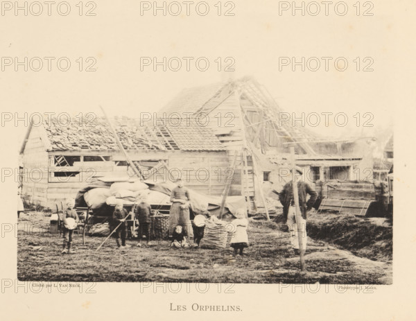 Children who were orphaned after an explosion of a munitions factory in Oosterweel near Antwerp in 1889, Les orphelins, page, photomechanical print, number: '13', photographer: L. van Neck, Oosterweel, cliché maker: anonymous, Belgium, printer (person): J. Maes (possibly), Antwerp, 1889, printer (person), paper, collotype, photoprint, height 165 mm x width 230 mm, Image on page 13., orphans, Explosion of a munitions factory in Oosterweel in 1889, A faded black and white scene shows a group of people standing and sitting in front of a cluster of wooden buildings with partially missing roof tiles and exposed rafters, scaffolding and ladders leaning against the structures, piles of sacks and covered carts in the yard, a man standing with a long pole, several children holding bowls or standing near baskets, rough grassy ground underfoot, and various wooden crates, fences, and farm equipment scattered around.