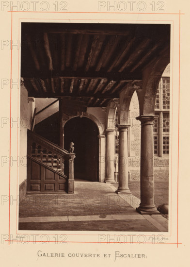 Covered corridor with stairs of the house of Christoffel Plantijn, Galerie couverte et escalier, page, photomechanical print, maker: Joseph Maes, Antwerp, c. 1873 - in or before 1878, paper, Woodburytype, height 164 mm x width 118 mm, corridor, staircase, A sepia-toned photograph of a covered gallery with a wooden beamed ceiling and a tiled floor, showing a carved wooden staircase with a decorative newel post on the left and a series of stone columns and arches on the right, sunlight casting shadows across the floor and the column bases, a dark arched opening in the background leading into shadow, a portion of a multi-paned window visible through one arch, and masonry walls with textured stonework.