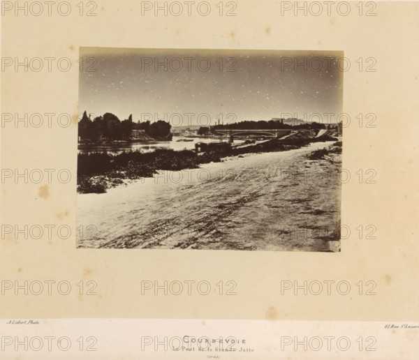 View of Courbevoie, Courbevoie. Le Pont de la Grande Jatte, photograph, page, number: '(No. 65)', address: '31, Rue St. Lazare', photographer: Alphonse-Justin Liébert, Courbevoie, 1871 - 1872, paper, albumen print, albumendruk, height 99 mm x width 130 mm, prospect of village, silhouette of village, Paris Commune (1871), A sepia-toned landscape photograph mounted on a larger beige matte shows a riverside scene with a wide path or shoreline in the foreground, textured ground leading toward the water, low vegetation and dark shrubbery along the riverbank, a calm river reflecting light, a bridge spanning the river with an arched design, clusters of trees and taller foliage on the opposite bank, buildings and low industrial structures partially visible behind the trees, a distant flat-topped hill or plateau on the horizon, and a sky mottled with light speckles.