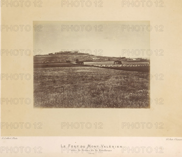 View of the fortress of Mont-Valérien in Suresnes, Le Fort du Mont Valérien avec la Ferme de la Fouilleuse, photograph, page, number: '(No. 72)', address: '31, Rue St. Lazare', photographer: Alphonse-Justin Liébert, Suresnes, 1871 - 1872, paper, albumen print, height 99 mm x width 132 mm, fortress, Paris Commune (1871), Mont Valérien, A sepia-toned photograph mounted on a wide beige matte shows a distant hilltop with a cluster of rectangular buildings and lower structures along its crest, a patchwork of fields and terraces spread across the slope and foreground, a few isolated trees and small groves dotting the midground, rows of small conical haystacks visible in one field, and a broad expanse of textured grassy land filling the lower portion of the image under a pale, lightly speckled sky.