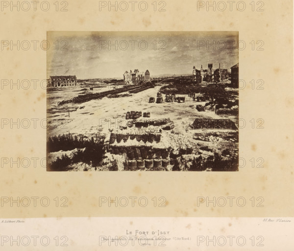 Le Fort d'Issy. Vue Générale du Panorama intérieur (Côté Nord), View of Fort d'Issy in Issy-les-Moulineaux after the destruction by the Paris Commune, photograph, page, number: '(No. 73)', address: '31, Rue St. Lazare', photographer: Alphonse-Justin Liébert, Issy-les-Moulineaux, 1871 - 1872, paper, albumen print, height 99 mm x width 136 mm, ruin of a building, architecture, fortress, Paris Commune (1871), Fort d'Issy, A sepia-toned photograph shows a wide open area with rows of stacked objects and scattered barrels in the foreground, leading to a rough terrain with pathways and tracks; several wheeled carts and more barrels are positioned further back; on the left and right sides of the midground there are ruined multi-story buildings with damaged roofs and exposed walls; the sky above is cloudy with a grainy texture.
