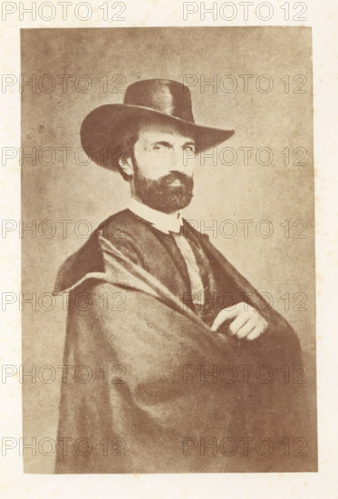 Portrait of Antoine Wiertz, photograph, page, check stamp: 'SOCIÉTÉ ROYALE BELGE DE PHOTOGRAPHIE, 69, RUE KEYENVELD, BRUXELLES', photographer: Edmond Fierlants, c. 1863 - in or before 1865, paper, albumen print, height 161 mm x width 106 mm, Person, Antoine Joseph Wiertz, historical persons, portrait, self-portrait of painter, A sepia-toned portrait of a bearded man wearing a wide-brimmed hat and a dark cloak draped over his shoulders, showing his crossed arms and one hand resting near his chest; his face is turned slightly toward the viewer, with visible eyes and mustache, and the background is a plain textured surface.