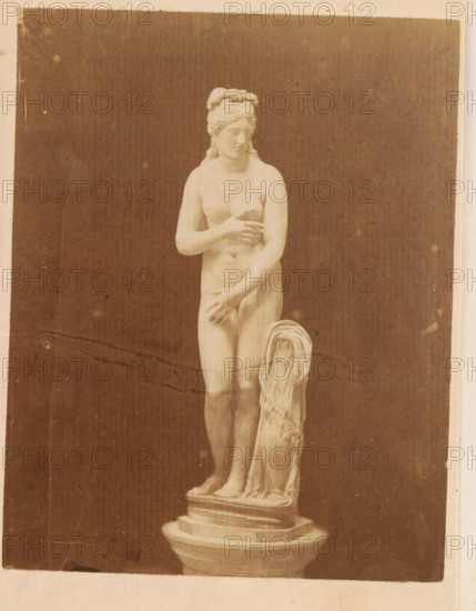 Sculpture of the Capitoline Venus in the Capitoline Museums in Rome, photograph, page, Capitoline Museums, c. 1872 - in or before 1882, photographer, Rome, paper, albumen print, height 128 mm x width 101 mm, Photo on frontispiece., sculpture, (story of) Venus (Aphrodite), A monochrome photograph of a marble statue of a nude female figure standing on a tiered round pedestal, with one arm bent across her chest and the other hand placed near her lower abdomen, her head turned slightly to one side with hair styled and tied at the back, a draped cloth hanging beside her and pooling at the pedestal, all set against a dark plain background with the photograph showing a light border and some surface marks and spots.