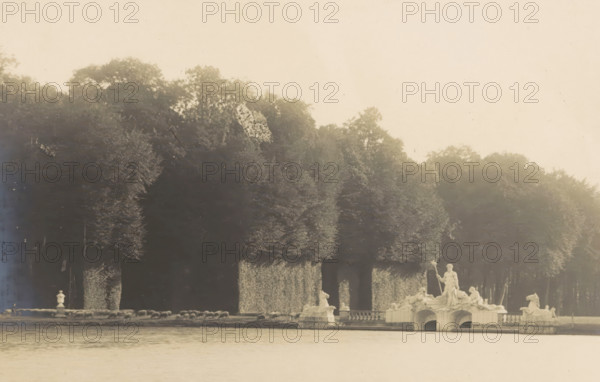 Le Lac et le Groupe de Neptune, View of a lake containing sculptures of Neptune, photograph, page, photographer: Coppin-Goisse, c. 1904 - in or before 1909, paper, gelatin silver print, developing gelatin silver print, height 83 mm x width 126 mm, sculpture, lake, Beloeil, A wide rectangular photograph bordered by a lighter mat shows a body of water in the foreground with a sculptural fountain group on a stone platform near the shore, featuring multiple classical figures and reclining horse forms, and a standing central figure holding a staff. Behind the fountain a row of closely planted, tall, trimmed trees forms a dense leafy screen with sculpted archways or openings at their bases. The sky above the trees is pale and largely empty, and the photograph is set against a brown textured backing with a thin painted rectangular border near its edges.