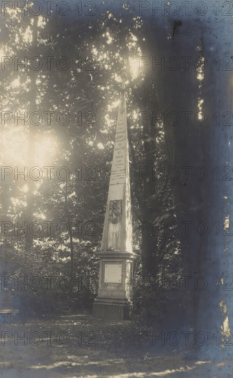 Obelisk of Prince Charles between trees, Obélisque du Prince Charles, photograph, page, photographer: Coppin-Goisse, c. 1904 - in or before 1909, paper, gelatin silver print, developing gelatin silver print, height 128 mm x width 84 mm, trees, obelisk, needle, Beloeil, A faded photograph mounted on a brown mat shows a tall stone obelisk standing on a stepped base surrounded by trees and foliage; sunlight filters through the leaves creating bright spots and reflections across the image, and the photo edges show signs of wear and aging.