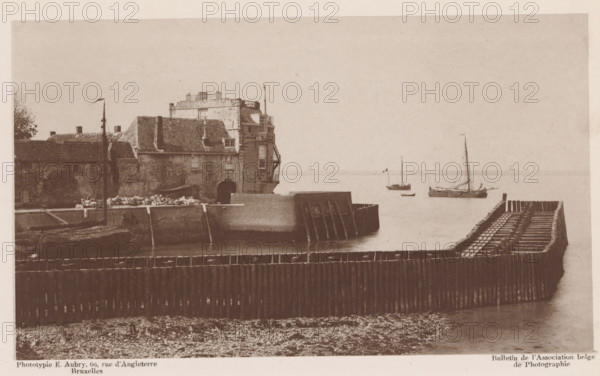 View of a dock, page, photomechanical print, inscription: 'Bulletin de l'Association belge de Photographie', inscription: 'Cliché au Gélatino-bromure', photographer: Hector Colard, cliché maker: Emile Aubry, 1878 - in or before 1883, Hector Colard, Emile Aubry, photographer, cliché maker, paper, collotype, photoprint, height 106 mm x width 179 mm, Print two pages before page 66., dry-dock, floating-dock, A sepia-toned harbor scene shows a weathered stone building with a tiled roof and visible chimneys situated on a quay, with an arched opening at the waterline and an external staircase on one side; wooden pilings and a curved timber breakwater extend into calm water in front of the building, and small sailing vessels with masts and rigging are moored a short distance offshore, one flying a small flag; the foreground shows a pebble-strewn shoreline and rows of vertical wooden posts forming a barrier, while the sky above is featureless and light in tone.