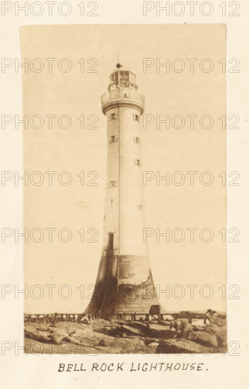 Bell Rock Lighthouse, View of the Bell Rock lighthouse, photograph, page, Scotland, c. 1867 - c. 1872, photographer, paper, albumen print, height 173 mm x width 119 mm, sea, lighthouse, lightship, A tall cylindrical lighthouse rising from a broadened base with visible texture and weathering, a narrow vertical seam running up its shaft, small rectangular windows set at intervals, a balcony with a railing near the top, and a glassed lantern room with a small finial above; rocky ground and a low wooden fence appear at the base, with a small structure near the lighthouse and scattered stones in the foreground.