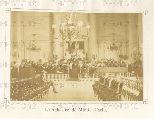 Les jeudis de Monte Carlo : impressions musicales, Ch.M. Domergue, book, dedication: 'à mon... le C... Barreme, souvenir ... de l'author, Domergue', date: 'JAN, 2003', bookplate: 'BIBLIOTHÈQUE du CHATEAU de BEAUREGARD', writer (clerical worker): Charles Mathieu Domergue, 1875, Charles Mathieu Domergue, writer (clerical worker), paper, printing, albumen print, height 188 mm x width 108 mm x thickness 20 mm, A sepia-toned photograph of an ornate interior showing rows of upholstered chairs arranged along a central aisle leading to a raised platform where a group of musicians are seated and standing with instruments, a conductor visible near the front and a large string instrument resting at the edge of the platform; the room features tall columns, decorative wall panels, multiple chandeliers hanging from the ceiling, wall-mounted candelabra, and framed artwork or mirror behind the musicians, with a single seated man visible in the aisle near the foreground.