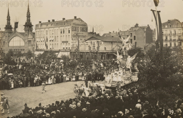 Wagon, glorification of Antoon van Dijck, photograph, page, Antwerp, c. 1899, photographer, paper, printing, height 98 mm x width 152 mm, civic architecture, edifices, dwellings, animal traction, animal-drawn vehicle (carriage etc.), festivities (decorated float, wagon, festive activities), A crowded street scene with a large procession float pulled by teams of horses and adorned with statues and figures, surrounded by a dense crowd of people wearing period clothing and hats, buildings with ornate facades and tall spires in the background, trees partially framing the scene, and lamp posts and banners visible above the assembled spectators.
