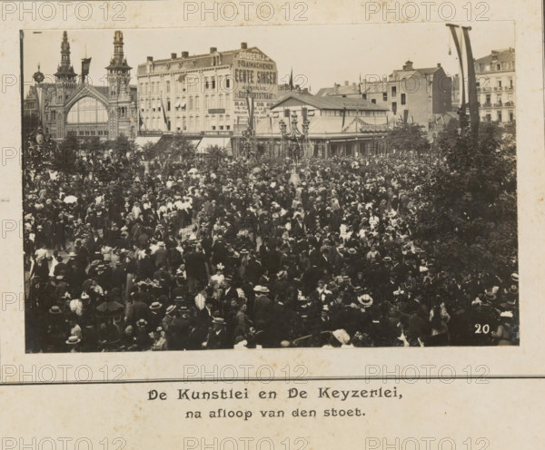 The Kunstlei and the Keyzerlei, after the procession, photograph, page, Antwerp, c. 1899, photographer, paper, printing, height 98 mm x width 152 mm, civic architecture, edifices, dwellings, festivities (decorated float, wagon, festive activities), A sepia-toned photograph showing a densely packed outdoor crowd with people wearing period clothing and hats, occupying a square in front of a row of ornate multi-story buildings with decorative facades and tall towers, a large arched window in one building, and a covered pavilion with a pitched roof. A tall ornate street lamp stands amid the crowd, trees or large bushes frame parts of the scene, and flags or long banners hang from poles near the buildings. The crowd includes men and women in dark and light garments, many holding parasols or wearing wide-brimmed hats, with groups clustered closely together across the foreground and middle distance.