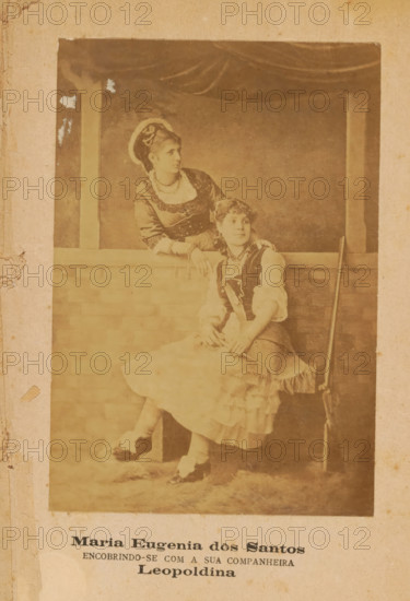 Portrait of Maria Eugenia dos Santos and an unknown woman, Maria Eugenia dos Santos, photograph, page, inscription: 'ENCOBRINDO-SE COM A SUA COMPANHEIRA, Leopoldina', c. 1881 - in or before 1886, photographer, paper, albumen print, height 140 mm x width 96 mm, Photo after cover page., Person, historical persons - BB - woman, A faded sepia photograph mounted on aged paper shows two women in period costumes posed by a low brick wall and a curtained backdrop. One woman stands behind the wall, leaning on it with one arm and looking to the side; she wears a dark bodice with decorative trim, a light blouse with elbow-length sleeves, a necklace, and a headpiece with a circular halo-like element. The other woman sits on the wall edge, facing slightly forward with hands resting in her lap; she wears a layered skirt with ruffled hem, a vest over a blouse, stockings, and shoes, and a necklace. A long slender object rests upright against the wall beside the seated woman. The photograph has worn ed...