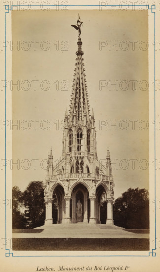 View of the Monument to the Dynasty, Laeken. monument du Roi Léopold 1er, photograph, page, Laeken, c. 1875 - c. 1880, photographer, Brussels, paper, albumen print, albumendruk, height 172 mm x width 98 mm, monument, statue, A tall ornate stone monument with a pointed spire and intricate Gothic detailing stands on a stepped base surrounded by trees, the lower section featuring arched openings supported by columns and a statue visible inside one of the arches, the spire topped with a sculpted figure and decorative finials, all set against a plain sky and framed by a light border.