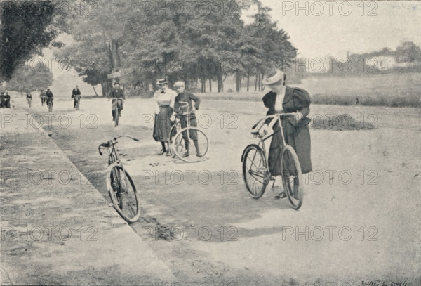 Au Bois de Boulogne, Cyclists in the Bois de Boulogne in Paris, France, page, photomechanical print, inscription: 'Agrandissement d'un cliché 6 1, 2x9 obtenu avec la Jumelle photographique de H. Mackenstein., Cet agrandissement a été fait avec la Jumelle elle-même adaptée sur l'agrandisseur économique Mackenstein., Voir l'épreuve 6 1, 2x9, page 300., Papier &lt, Perfection &gt, qualité A. - Grosvenor, Chater et Cie, 6, rue Favart, Paris.', address: 'Fabrique d'appareils pour la photographie. H. MACKENSTEIN, 15, rue des Carmes, Bois de Boulogne, cliché maker: Bonneau & Co., Geneva, c. 1891 - in or before 1896, Bonneau & Co., photographer, cliché maker, Paris, Genève, paper, autotype, similigravure, height 106 mm x width 156 mm, riding, cycling (recreation), park, A faded black-and-white scene shows a wide tree-lined pathway with several bicycles and people. In the foreground a woman in a dark dress and a hat stands holding a bicycle, looking down toward the ground. Nearby a bicycle lean...