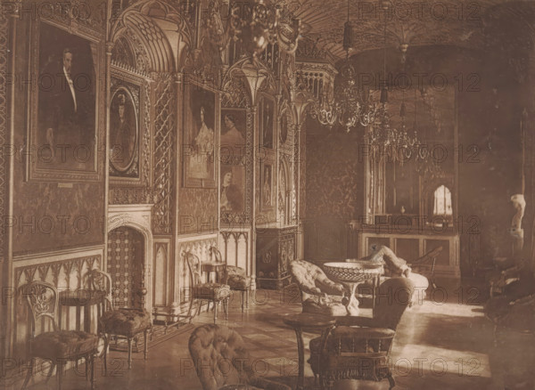 Interior of the picture gallery in Strawberry Hill in London, page, photomechanical print, photographer: Ernest Edwards, Strawberry Hill, 1869 - in or before 1871, Twickenham, paper, collotype, photoprint, height 163 mm x width 222mm, art collection, art gallery, The image shows an ornate interior room with carved wooden paneling and arched decorative niches along one wall, each niche containing framed portrait paintings. Upholstered chairs and small round tables are arranged in the foreground and midground, including tufted armchairs and stools with carved legs. A carved fireplace surround and patterned wall coverings are visible beneath the portraits. Elaborate chandeliers hang from a vaulted, decorated ceiling. In the background there is a raised area with a railing or counter and a small arched window opening, and a sculpted figure or statue is positioned near the far right wall. Flooring with a subtle geometric pattern stretches across the space.