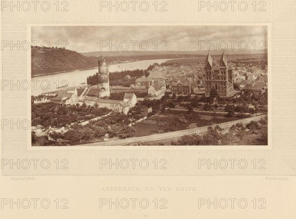 Andernach, on the Rhine, View of Andernach on the Rhine, page, photomechanical print, inscription: 'Europe', photographer: William England, cliché maker: Woodbury Permanent Photographic Printing Company, c. 1870 - in or before 1875, Rhineland-Palatinate, paper, pasteboard, Woodburytype, Woodburytypie, height 83 mm x width 163 mm, city-view in general, 'veduta', river, Rhine, A sepia-toned panoramic view shows a riverside town with a wide waterway running horizontally across the scene and a terraced hillside on the far bank. In the foreground are gardens and orchards with small pathways and low walls, and a road running along the bottom edge. Clustered houses with pitched roofs fill the middle ground, interspersed with taller buildings and rows of trees. A cylindrical stone tower with a battlemented top stands near the river, and a large stone church with twin tall spires and ornate façades rises above surrounding buildings on the right side. Small boats are visible on the river, and th...