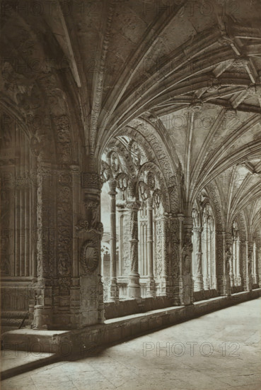 The cloisters of Belem, Cloister of the Hieronymite Monastery in Lisbon, page, photomechanical print, inscription: 'Stuart photo.', inscription: 'Europe', photographer: unknown, Mosteiro dos Jerónimos, cliché maker: Woodbury Permanent Photographic Printing Company, c. 1870 - in or before 1875, Lisbon, paper, pasteboard, Woodburytype, Woodburytypie, height 158 mm x width 106 mm, cloisters, monastery, A sepia-toned photograph shows a long covered walkway with a vaulted ceiling supported by a series of ornate stone arches and carved columns. The ceiling features ribbed vaulting with decorative bosses at the intersections, while the arches and column surfaces display intricate relief carvings of foliage and geometric patterns. Light falls through tall arched openings along one side, casting soft shadows on the smooth stone floor and the raised stone ledge that runs beside the columns. The photograph is mounted on a textured card with a patterned border surrounding the image.