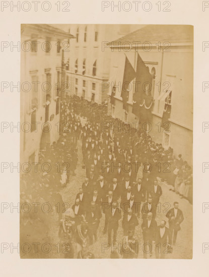 Funeral procession of Henri d'Artois, photograph, page, photographer: A. Vallois, Göritz (Austria), c. 1878 - in or before 1883, A. Vallois, photographer, Steiermark, paper, albumen print, height 211 mm x width 155 mm, cortege, funeral procession, A sepia-toned photograph showing a narrow street crowded with people in formal dark suits and bow ties walking in an organized procession, flanked by onlookers along the sides and leaning from windows of adjacent multi-story buildings, with large dark flags draped from windows on one side and decorative swags hanging beneath some windows, cobblestone pavement underfoot, and ornate uniforms and helmets visible on a few figures near the front of the procession.