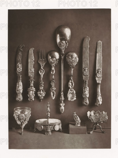 Twelve silver objects of the Polish royal house, displayed during the Exposition Rétrospective of 1873 in Tours, France, page, photomechanical print, maker: Gabriel Blaise, Tours, 1873, paper, Woodburytype, Woodburytypie, height 194 mm x width 141 mm, temporary exhibition, 'Salon', table-ware, Period, 1873-05-03 - 1873-05-25, A vertical photograph of ornate metalware arranged against a dark backdrop, showing a row of decorated utensils suspended near the upper half including several knives with elaborately carved handles and blades, a fork with a decorative stem, and multiple spoons with sculpted bowls and ornate handles; below them are decorative objects placed on a surface including a goblet with a raised relief design and tripod feet, a round footed dish with embossed sides and a central finial, a small sculptural figure mounted on a rectangular block, and a bowl with a textured, petal-like edge supported on a decorated tripod base.